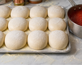 Pizza dough balls on a tray with a pot of tomato sauce in the background.