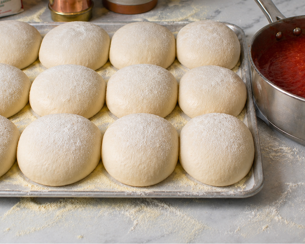 Pizza dough balls on a tray with a pot of tomato sauce in the background.